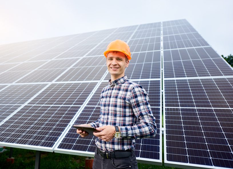 Smiling man in helmet with tablet in hands stands near solar panels on green plantation.